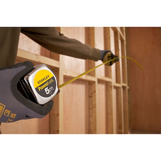 STANLEY 0-33-195 PowerLock 5m tape measure held in gloved hand, measuring wooden wall frame.