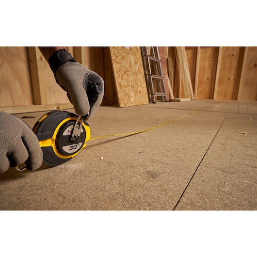 Photo of STANLEY 0-34-134 long tape measure in use on a construction floor, gloved hands holding the tool, side angle.