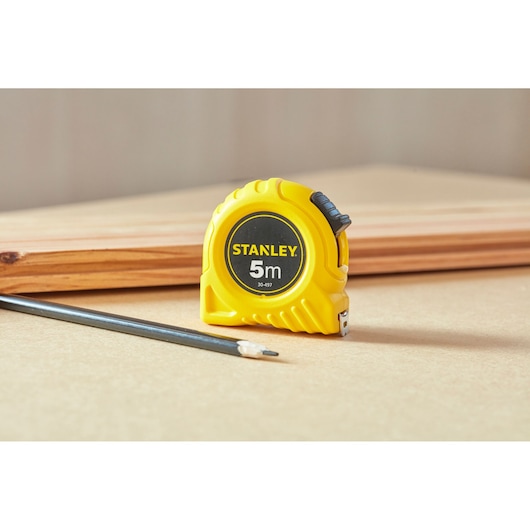 Stanley 1-30-497 tape measure in yellow casing, placed beside pencil and wood, product photography.