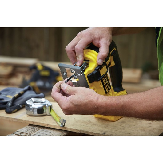 Photo of STANLEY FMC650M2 cordless jigsaw being adjusted by hand on a workbench with measuring tape and gloves.