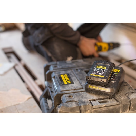 STANLEY FMC692L battery charger on tool case, close-up, worker using drill in background indoors.