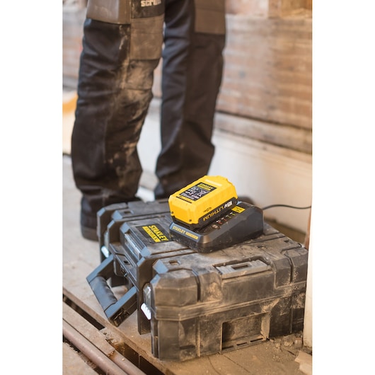 STANLEY FMC692L battery charger and yellow battery on rugged black toolbox, photo at worksite with person in background.