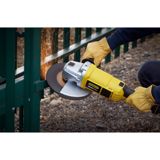 STANLEY FME841K angle grinder in use, cutting metal fence, yellow housing, worker wearing gloves.