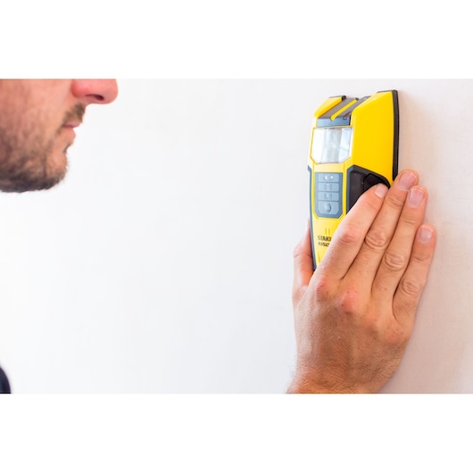 STANLEY FMHT0-77407 stud finder in yellow held against a white wall by a hand, front angle.