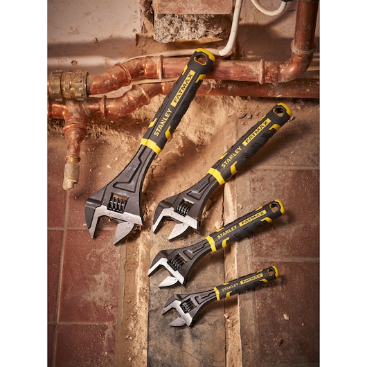 Four Stanley adjustable wrenches in different sizes placed on a tiled, dusty floor near copper pipes.
