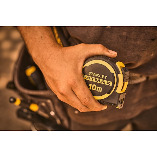 Photo of STANLEY FATMAX 10m tape measure FMHT33005-0 in hand, tool belt and tools in background.