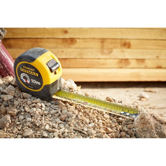 Photo of STANLEY FMHT36142-0 FATMAX 10m tape measure on rocky ground with yellow timber in background.