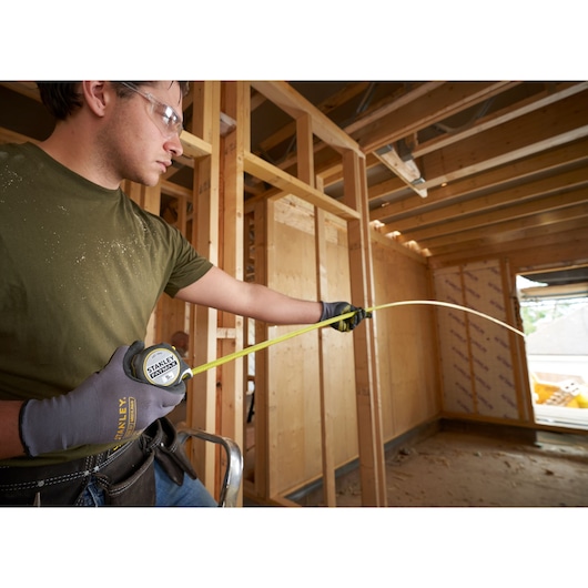 Stanley FMHT38214-0 tape measure used by worker in gloves at wood frame construction site.
