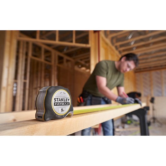 STANLEY FMHT38214-0 FATMAX tape measure on wood plank, construction scene with person in background.