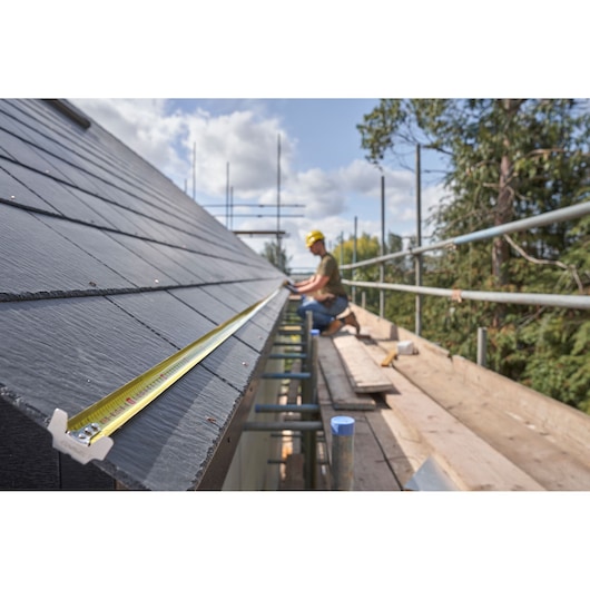 STANLEY FMHT38217-0 tape measure on roof edge, construction worker in background, outdoor setting.