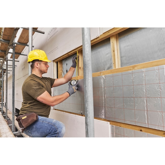 STANLEY FMHT38217-0 tape measure used by worker in yellow hard hat, measuring insulation on construction site, side angle.