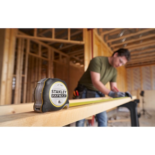 STANLEY FMHT38217-0 tape measure on a wood beam, with worker using it at a construction site.