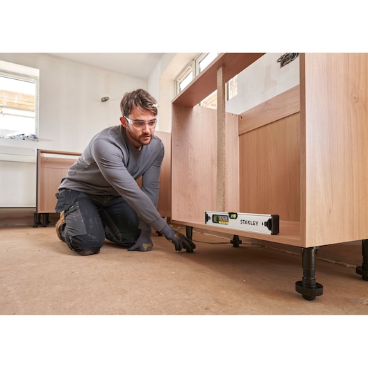 Stanley FMHT43670-1 spirit level placed under a wooden cabinet during installation, with worker adjusting cabinet legs.