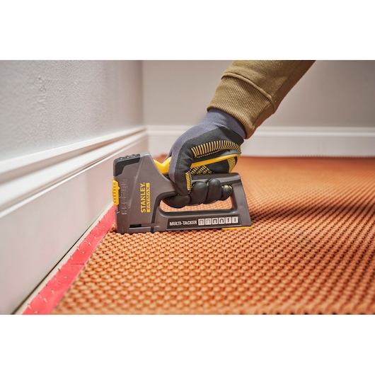 Stanley FMHT6-70868 multi-tacker stapler in use, held by gloved hand installing carpet near wall.