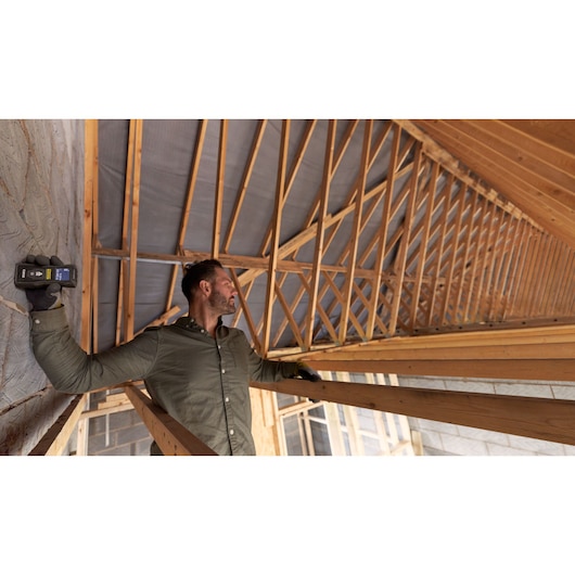 STANLEY FMHT77165-0 laser measuring tool used by worker in attic with wooden roof beams.