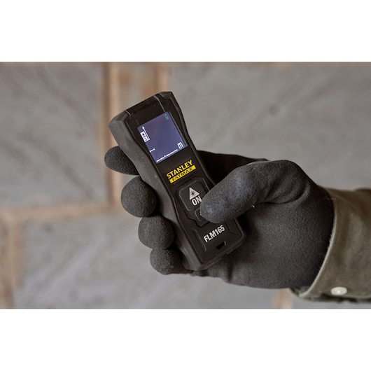 Photo of STANLEY FMHT77165-0 laser distance measurer held in a gloved hand, screen and buttons visible.