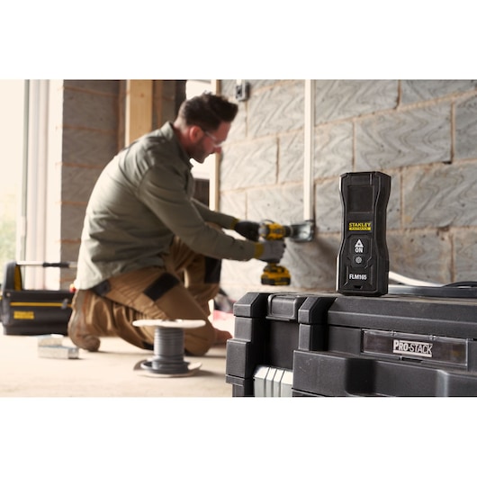 STANLEY FMHT77165-0 laser measure in foreground, on PROSTACK case, worker drilling in background.