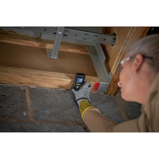 STANLEY FMHT77165-0 laser distance measurer in use on wooden beam, user wearing safety gloves.