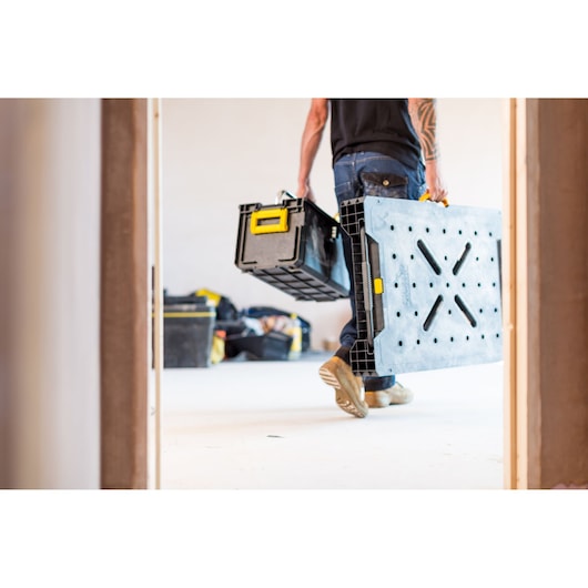 STANLEY FMST1-75672 portable folding workbench carried by person, black with yellow accents, indoor setting.