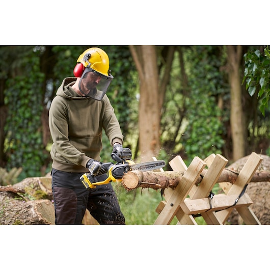 STANLEY SFMCCS730B chainsaw in use, person cutting a log outdoors on a wooden sawhorse, forest background.