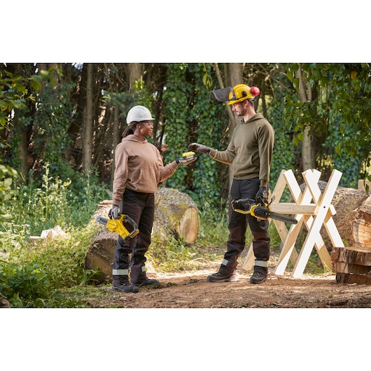 STANLEY SFMCCS730B chainsaw held by two people outdoors, with safety gear, near logs and sawhorse.