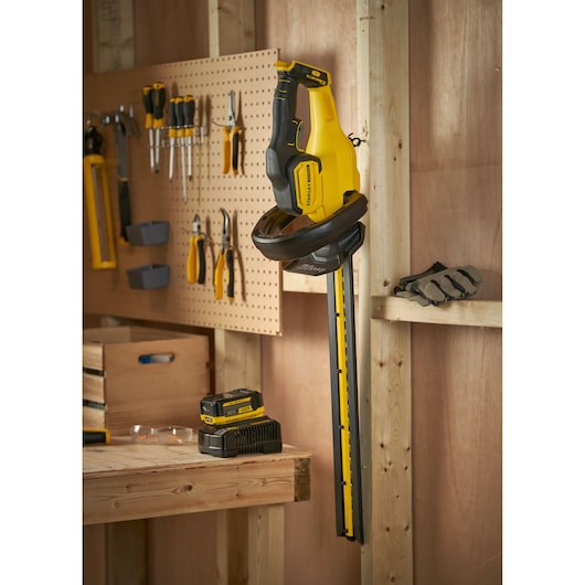 Photo of STANLEY SFMCHT655B hedge trimmer hanging in a workshop with tools and battery charger visible.