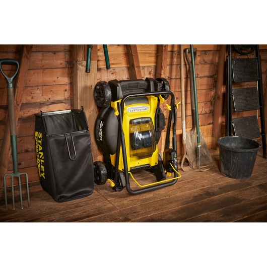 STANLEY SFMCMW2651M lawn mower shown folded upright next to black grass collection bag in a wooden shed.