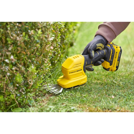 STANLEY STCSS20B hedge grass shear being used to trim grass at the edge of a bush, close-up outdoor view.