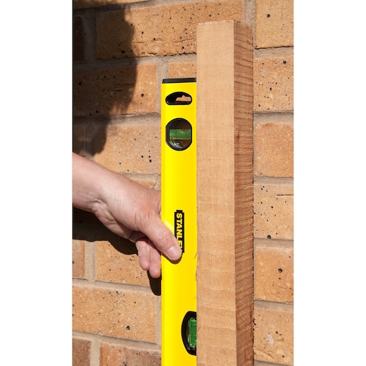 STANLEY STHT1-43103 yellow spirit level held against wood and brick wall, close-up view.