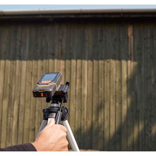 STANLEY STHT1-77139 laser distance measurer on tripod held by hand, outdoor setting with wooden wall.