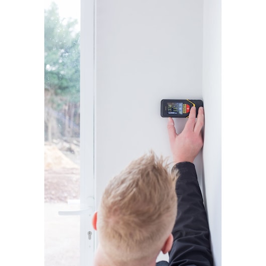 STANLEY STHT1-77140 laser distance measurer held against white wall by person, side angle.