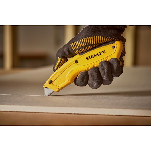 Photo of STANLEY STHT10430-0 utility knife cutting drywall, held by gloved hand.