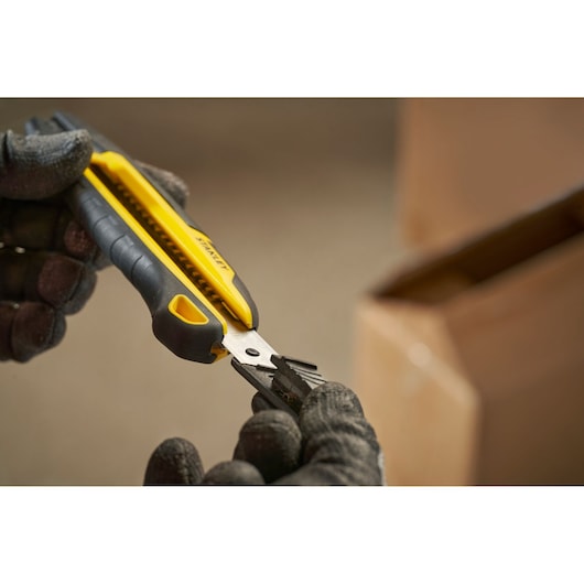 Stanley STHT10501-0 utility knife, yellow and black, held in gloved hands beside a cardboard box.
