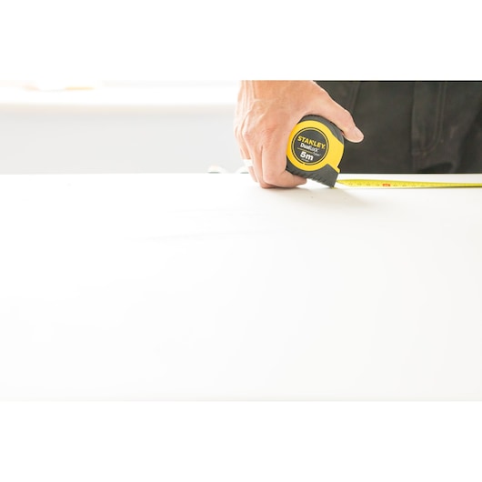 Photo of STANLEY STHT36803-0 tape measure held by hand, yellow and black casing, measuring on white surface.