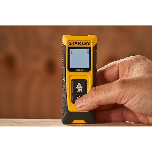 Photo of STANLEY SLM65 laser distance measurer SKU STHT77065-0 in hand, yellow and black, display screen visible.
