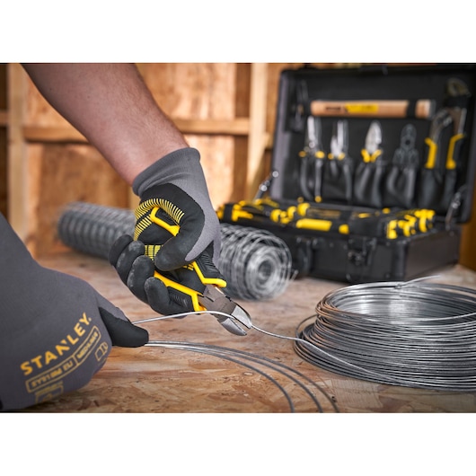 Photo of STANLEY STMT72986-1 pliers cutting wire, gloved hand holding tool, tool kit and wire rolls in background.