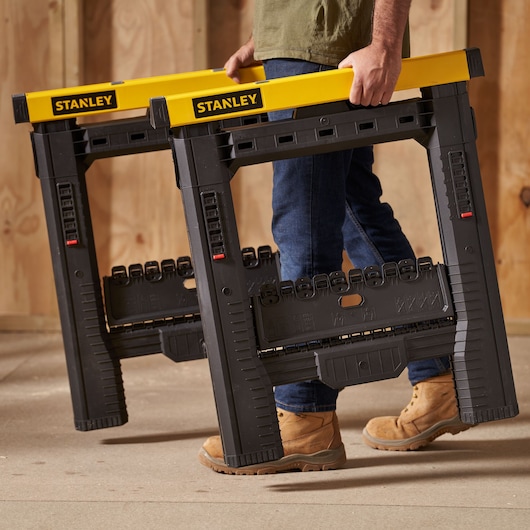 Photo of STANLEY STST1-70559 black and yellow folding sawhorses carried by a person in work boots indoors.