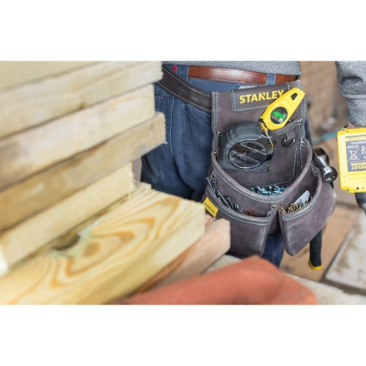 Photo of STANLEY STST1-80116 tool belt with tools and hardware, worn by a worker near stacked wooden planks.