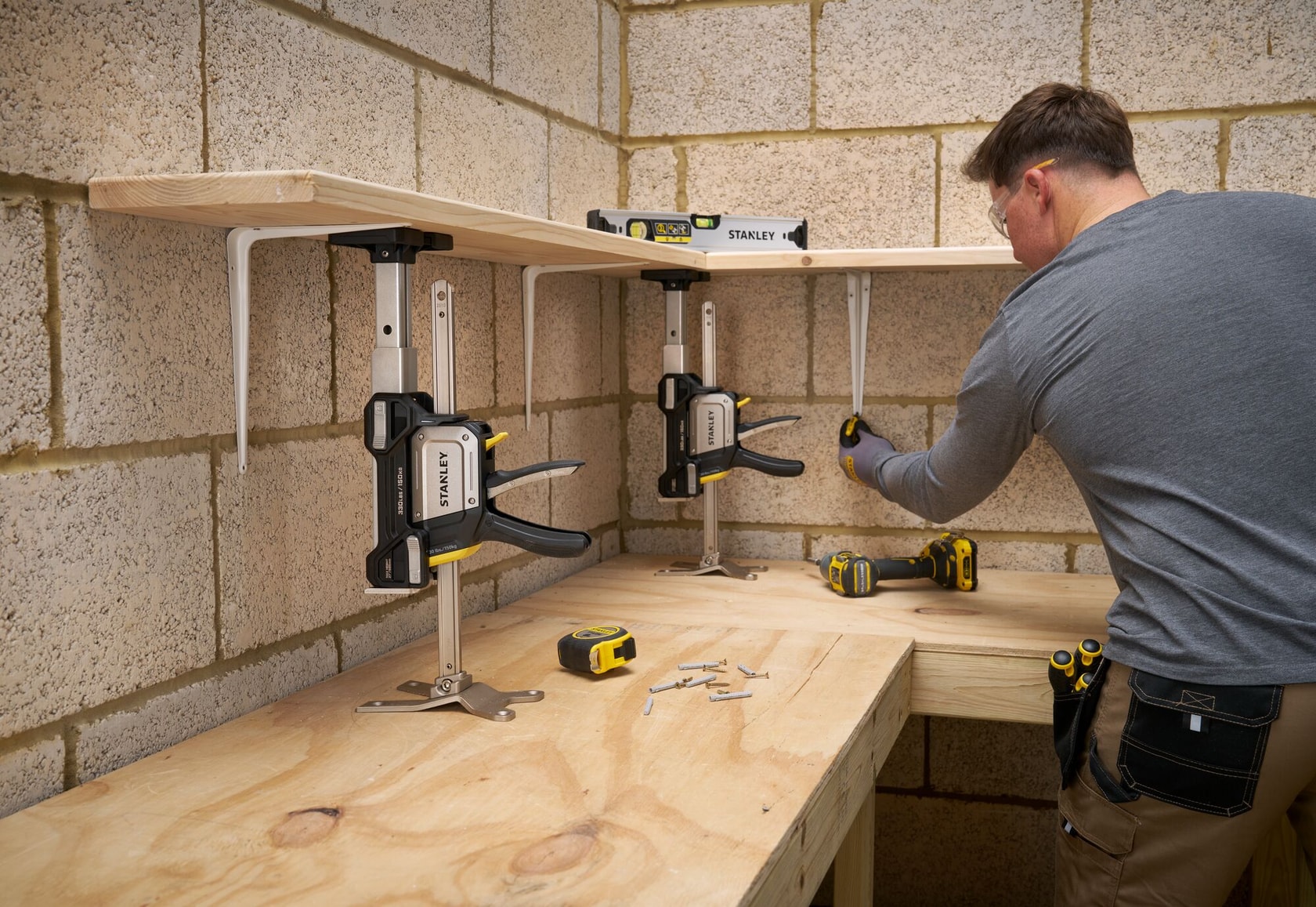 STANLEY FMHT83554-1 Zwinge hält Holzregal, Werkstatt mit Werkzeugen und Mann beim Zusammenbau, Frontansicht.